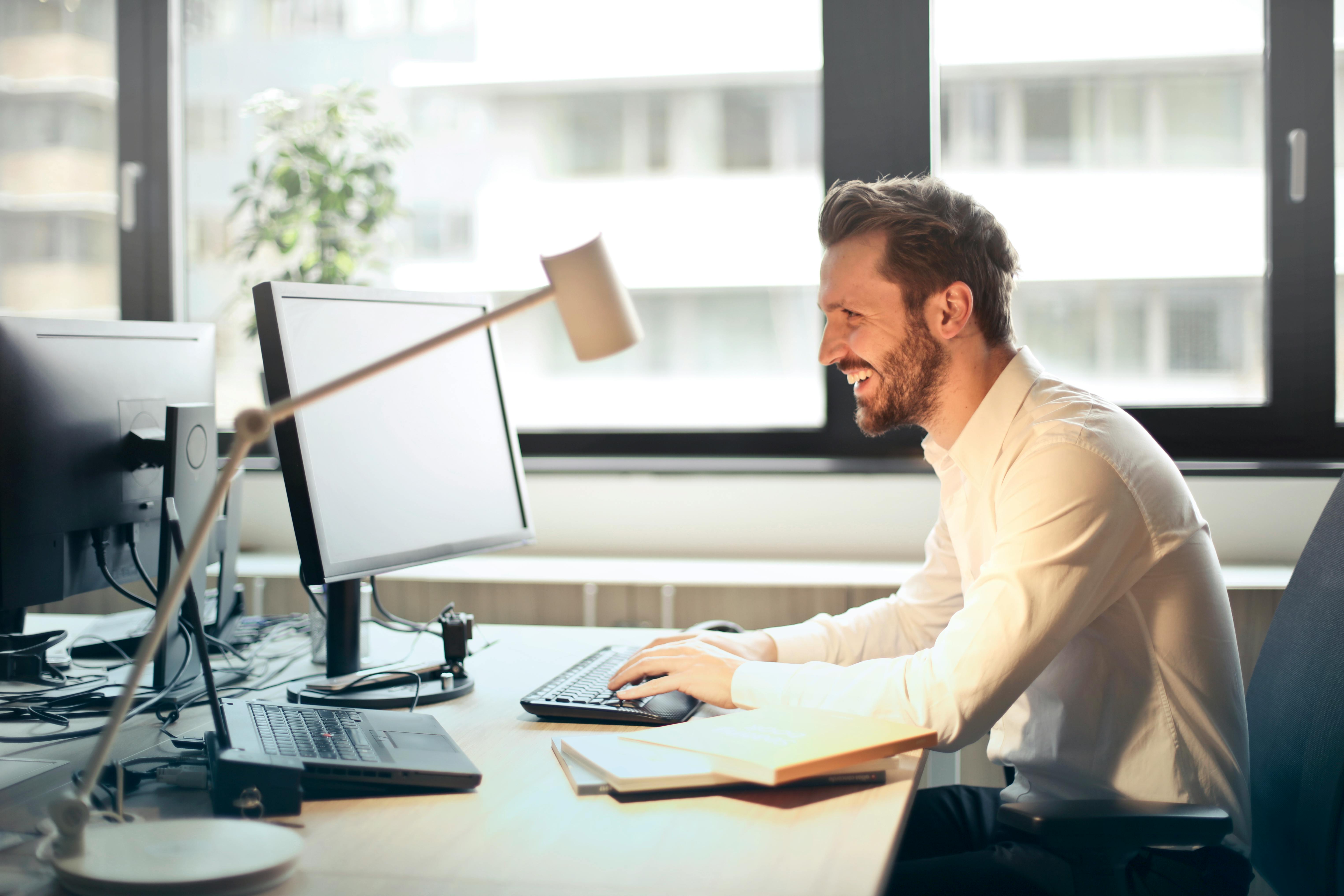 man in dress shirt smiling with typing on computer at a desk